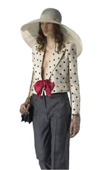 A model wearing a wide-brimmed white sun hat, a cream blazer with black polka dots and a large red bow at the waist, dark gray trousers, and holding a black clutch bag.