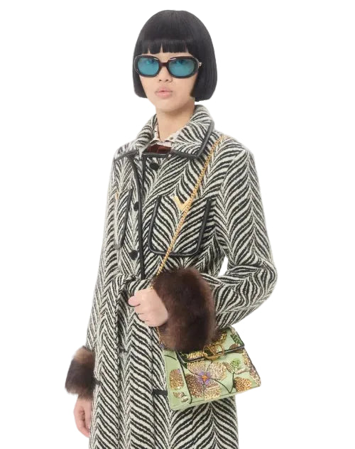 A model wearing a black and white patterned coat with a brown fur-trimmed collar, accessorized with a brown shoulder bag and a black hat.