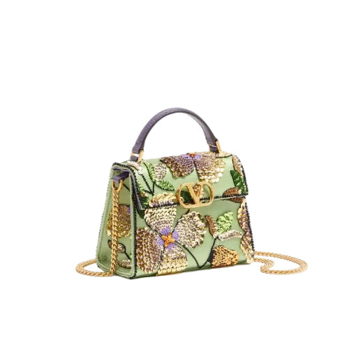 A structured green handbag with an intricate floral embroidery and beadwork pattern in gold, purple, and green, featuring a dark purple top handle, gold-tone hardware, and a gold chain shoulder strap, set against a plain white background.