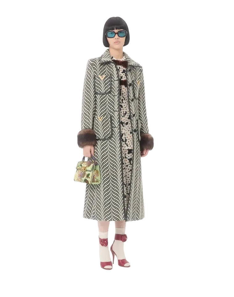 A model stands against a plain white studio background wearing a long patterned coat with dark fur trim, a black bob haircut, sunglasses, a patterned top, white socks, red heels, and holding a small patterned handbag.