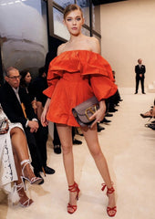 Model wearing a red off-shoulder dress on a fashion runway with an audience.