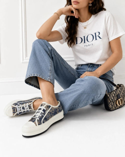 A woman sits on a white floor wearing a white Dior Paris graphic T-shirt, light blue jeans, and Dior monogram low-top sneakers. A small Dior monogram handbag rests beside her.